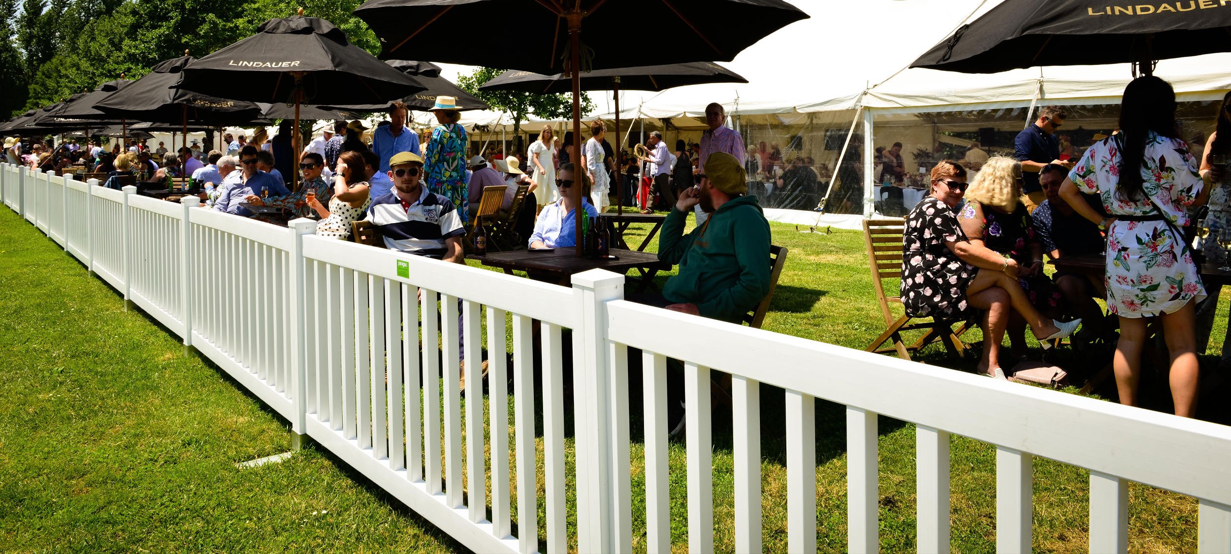 Temporary white PVC fencing at the races with dozens of outdoor dining tables with umbrellas giving everyone some shade