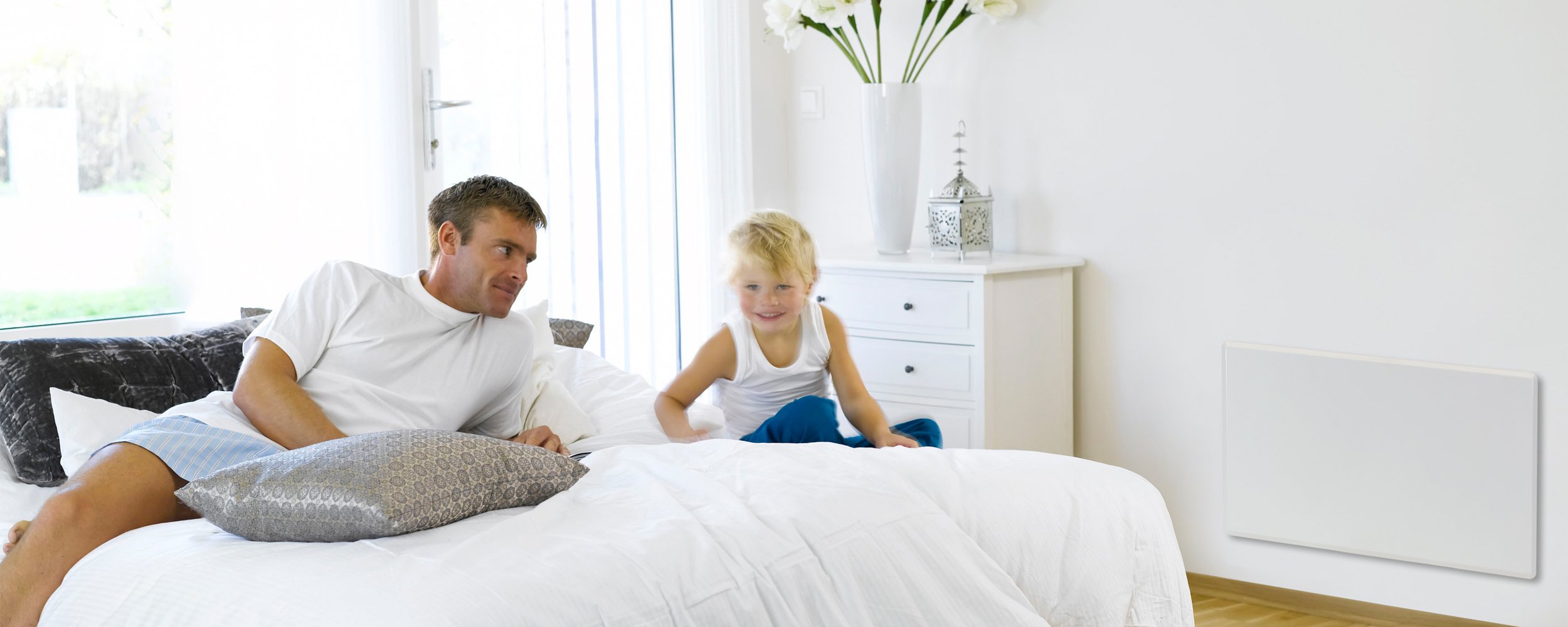 Father and young son sitting on a bed surrounded by white coloured furniture and an electric panel heater attached to the wall