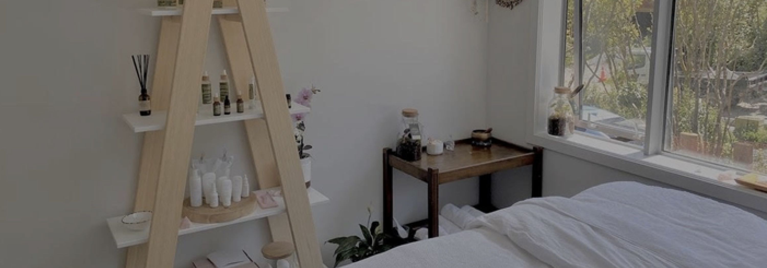 Spa bed with white sheets in a small white room with an open window and plenty of facial products on shelves