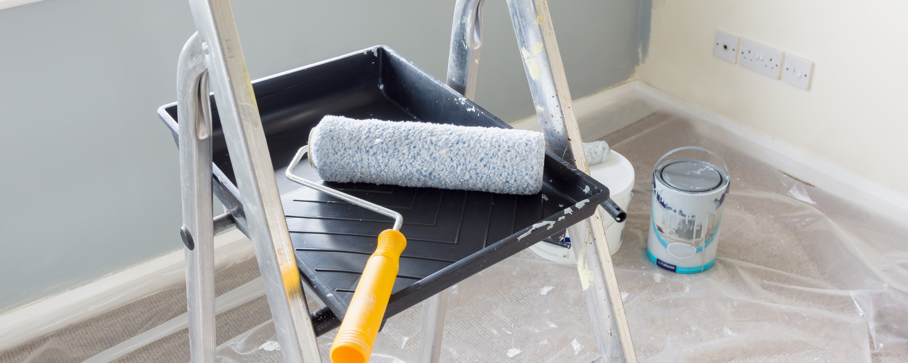 Yellow-handled paint roller sitting in a clean pain rolling tub on a ladder in a room with a half painted wall