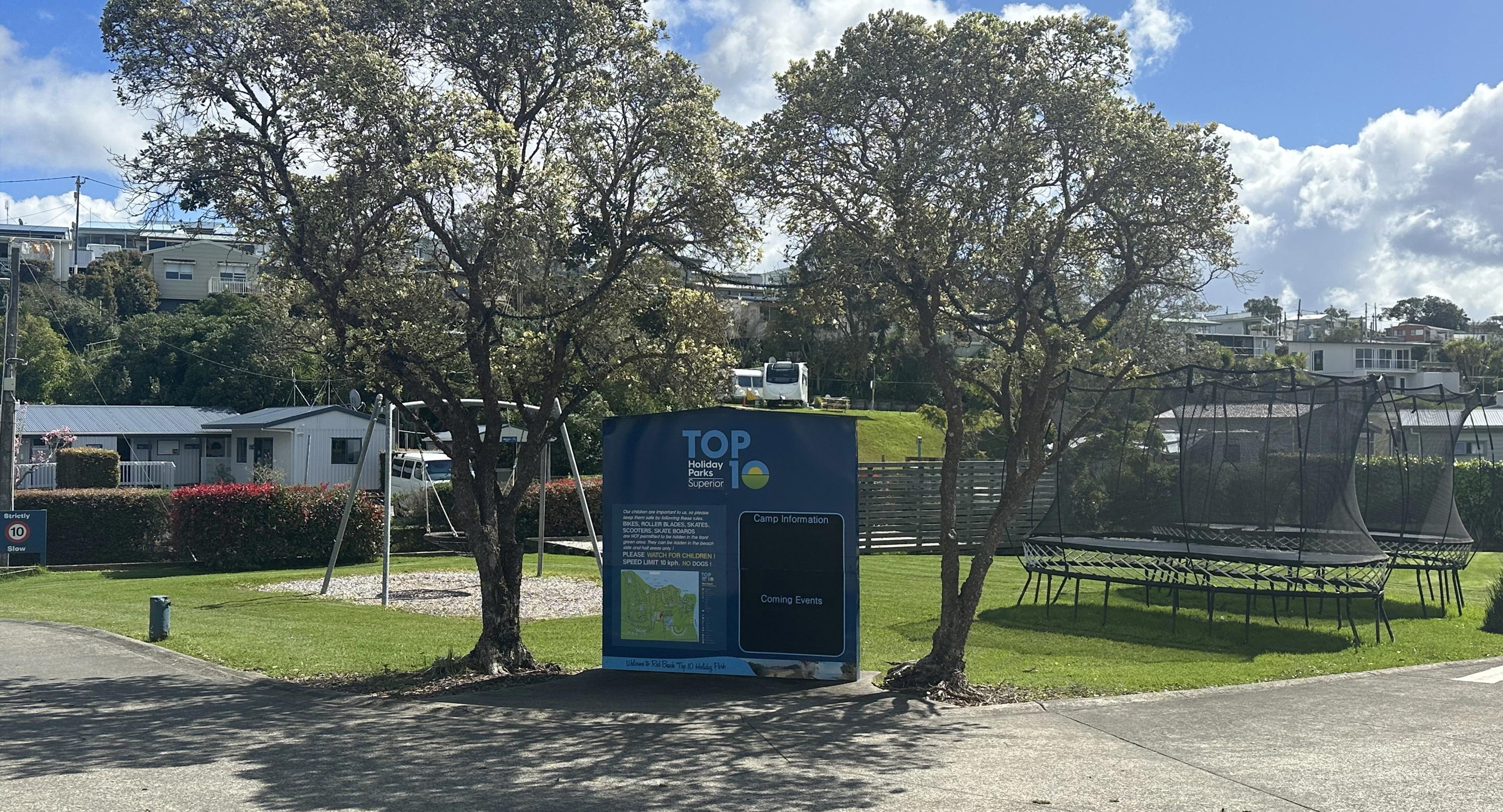 View of the Park sign and map at Pinewoods Holiday Park. A kids playground, swing-set and two large trampolines are pictured in the background