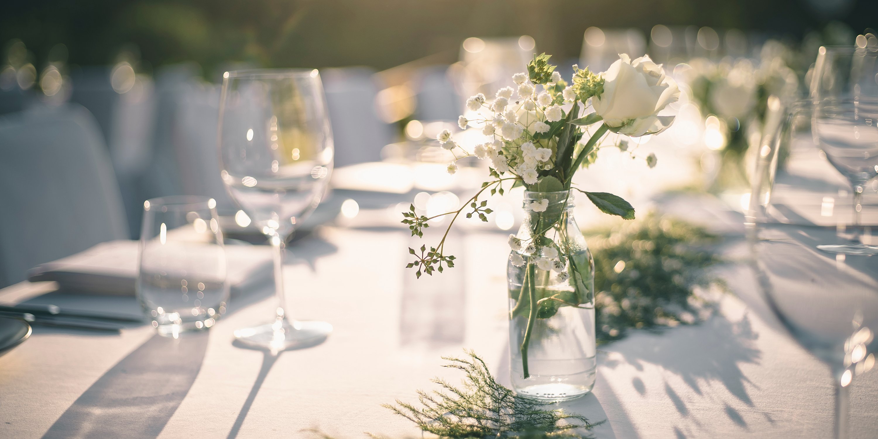 Close up view of some small white flowers in a vase on a table decorated with a white cloth and wine glasses in the sunset lighting | Party Hire Levin