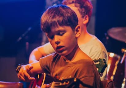 Little boy on stage with colourful lights as he plays the guitar with the rest of the band behind him