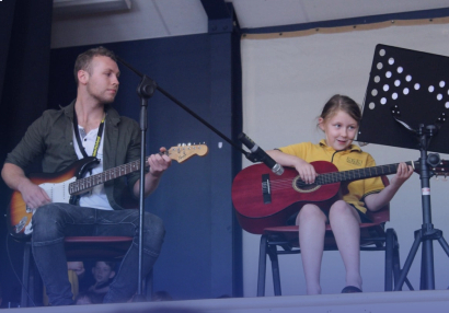 Music teacher and young student sitting on stage playing their guitars while the student reads the music
