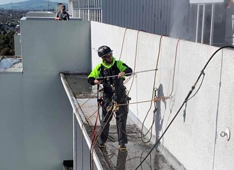 Commercial abseiler standing on the edge of a building's facade using a power washer to clean the building