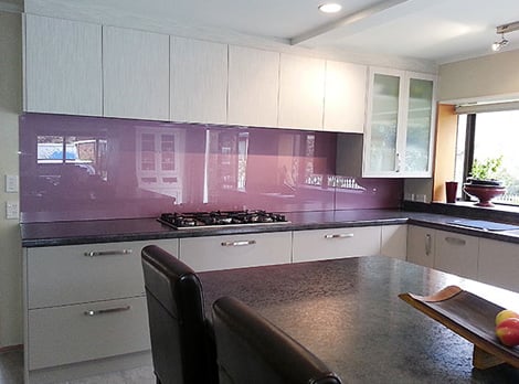 Kitchen with big off-white cabinetry, a dark grey benchtop with a large gas stove and a glossy purple splashback reflecting in the sunlight coming from the window