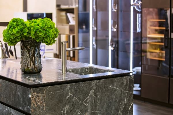Dark marble benchtop on a kitchen island with sleek silver fixtures and a bushy green plant with modern light strips in the background
