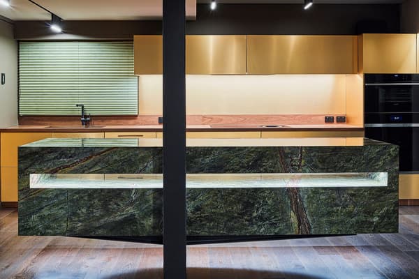 Green and blue nature-like granite island benchtop in a kitchen with goldish-orange wood cabinets and white lighting