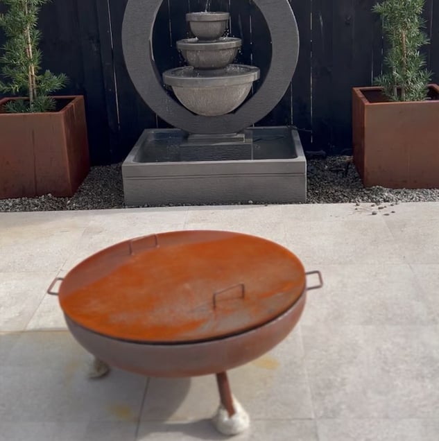 Outdoor firepit on a stone tile floor with two square flower pots and an articulate grey water fountain structure