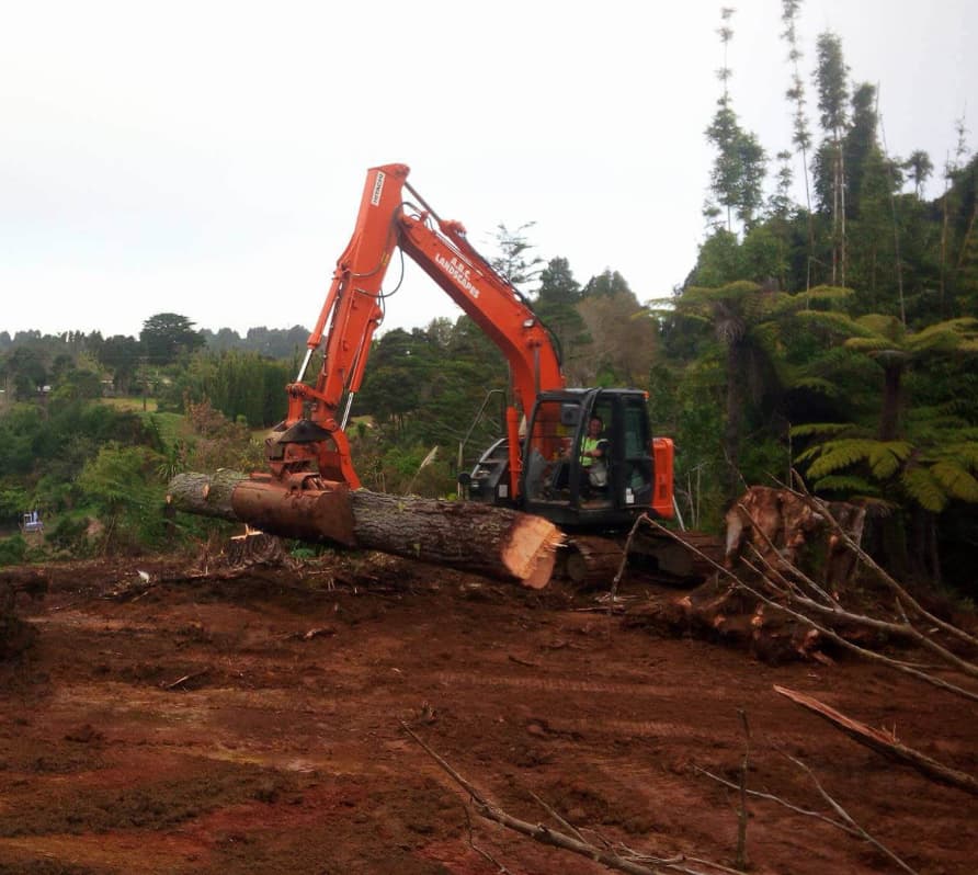 Large orange digger with a claw bucket lifting a massive piece of timber in the middle of a field surrounded by nature