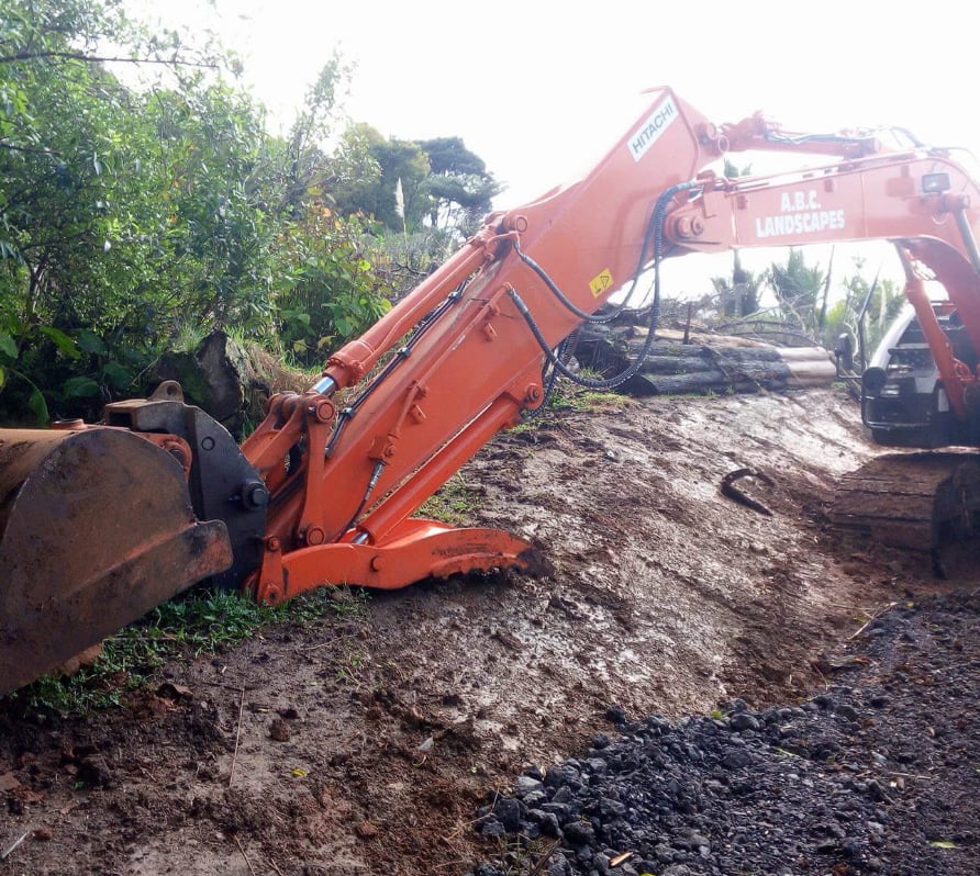 Large orange digger moving debris on a site with stacks of timber and vibrant green trees in the background